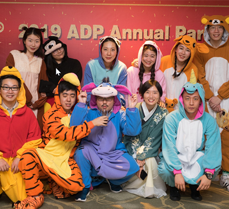 Men and women dressed in hooded animal onesie pajamas posing for a picture in front of a sign that reads 2019 ADP Annual Party