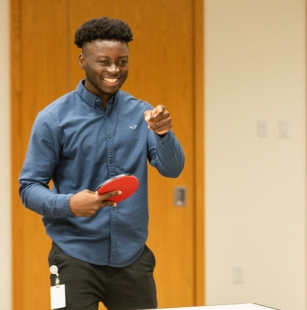 man holding ping pong paddle, smiling and pointing across table